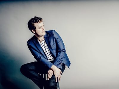 Benjamin Grosvenor in a blue blazer and striped shirt sitting on a stool against a neutral background, gazing to the side
