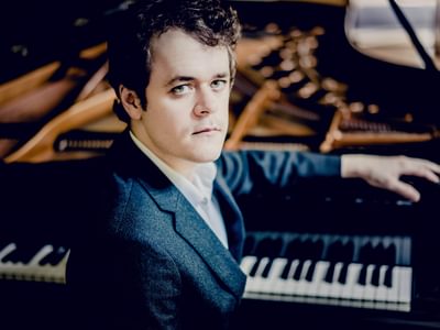 Benjamin Grosvenor in a blue suit, sitting at a grand piano, hand resting on the keys. He is gazing intently towards the camera
