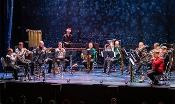 A group of musicians (Philharmonia) in festive attire playing brass instruments on stage. A person in a Santa hat conducts, with a starry backdrop