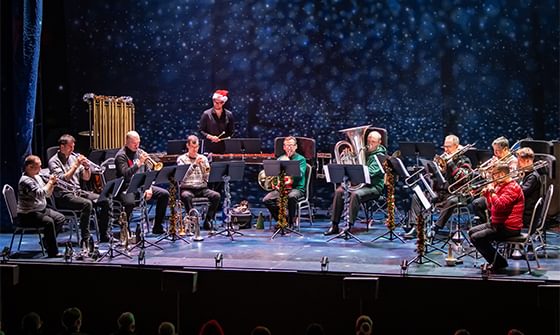 A group of musicians (Philharmonia) in festive attire playing brass instruments on stage. A person in a Santa hat conducts, with a starry backdrop