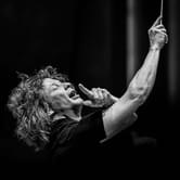 Black and white image of conductor Santtu-Matias Rouvali with curly hair, passionately leading an orchestra with his baton raised, eyes closed, and mouth open