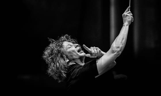 Black and white image of conductor Santtu-Matias Rouvali with curly hair, passionately leading an orchestra with his baton raised, eyes closed, and mouth open