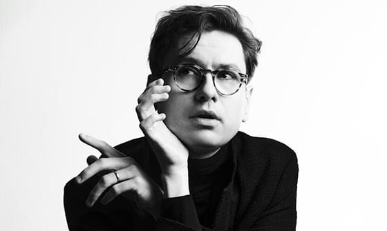 A black and white photo of Víkingur Ólafsson with glasses, looking pensive. He is resting his chin on one hand, raising the other in a gesture, wearing a dark outfit