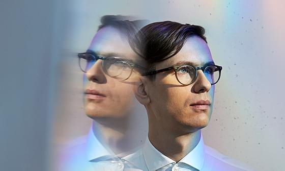 Víkingur Ólafsson wearing glasses and a white shirt is shown with a double exposure effect. His expression is thoughtful, and the image has a soft, rainbow-hued glow