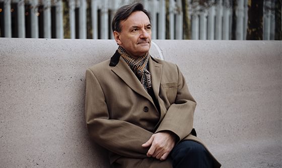 Sir Stephen Hough in a tan coat and patterned scarf sitting on a stone bench, looking into the distance