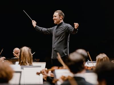 Conductor Roberto Gonzalez-Monjas in black attire energetically leads an orchestra, baton raised, with musicians focused on their instruments