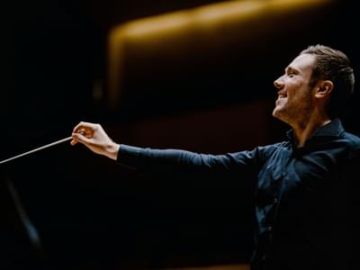 Conductor Roberto Gonzalez-Monjas wearing a dark shirt, leading an orchestra with a baton, smiling away from the camera