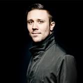 Roberto Gonzalez-Monjas wearing a dark jacket, standing against a black background. He is looking towards the camera with a calm expression