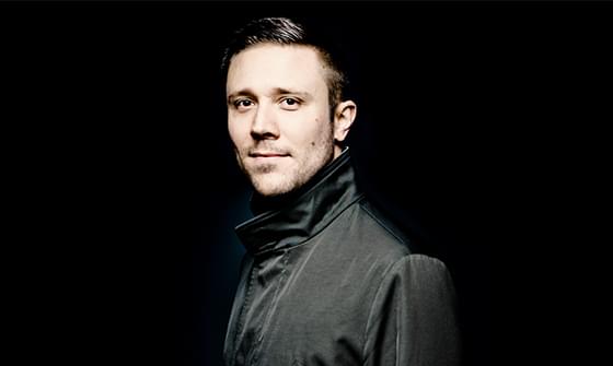 Roberto Gonzalez-Monjas wearing a dark jacket, standing against a black background. He is looking towards the camera with a calm expression