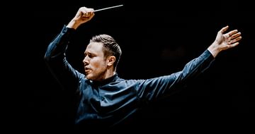 Roberto Gonzalez Monjas energetically directing an orchestra, one hand raised with a baton, the other outstretched, set against a black background