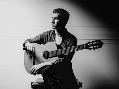 Black and white portrait of Thibaut Garcia seated, holding an acoustic guitar, gazing away from the camera