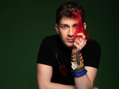 Thibaut Garcia with short hair leaning on a table against a green background. He is holding a red feather and wearing bracelets