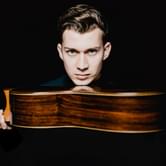 Thibaut Garcia, a man with short dark hair gazing intensely at the camera, holding a wooden guitar horizontally with his chin resting on it, on a black background
