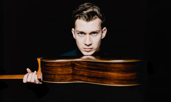 Thibaut Garcia, a man with short dark hair gazing intensely at the camera, holding a wooden guitar horizontally with his chin resting on it, on a black background
