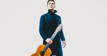 Thibaut Garcia, a man in a dark coat standing and holding a classical guitar. He is looking up against a light background