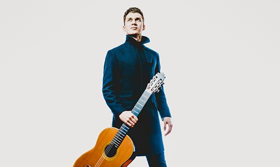 Thibaut Garcia, a man in a dark coat standing and holding a classical guitar. He is looking up against a light background