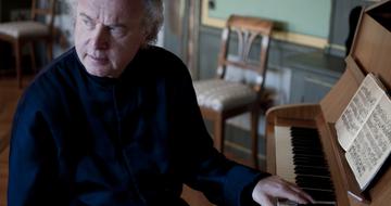 Sir András Schiff at his piano looking away from it and with one hand on the keys