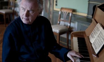 Sir András Schiff at his piano looking away from it and with one hand on the keys