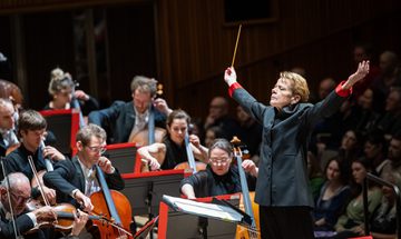 Marin Alsop, an older white woman with short light blonde hair in front of an orchestra conducting in a black suit with red cuffs