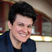 Aziz Shokhakimov, a young white man with quaffed black hair in a blue blazer smiling at the camera