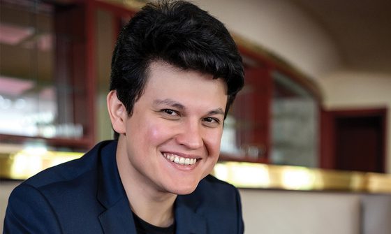 Aziz Shokhakimov, a young white man with quaffed black hair in a blue blazer smiling at the camera
