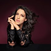 Beatrice Rana, an Italian woman with dark curly hair smirking and leaning her head on her hands in a black lace blouse against a dark reddish purple background