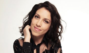 Beatrice Rana, an Italian woman with dark curly hair smirking with one hand by her chin in a black lace blouse against a white background