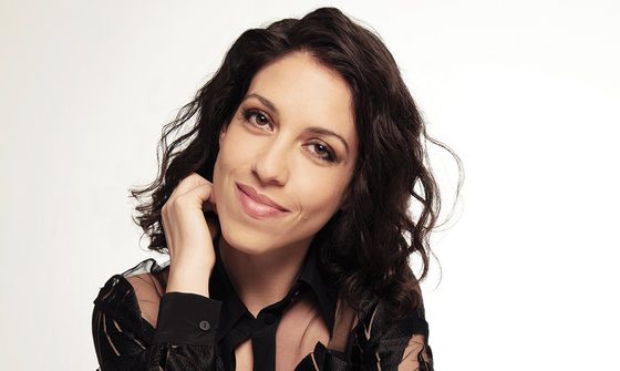 Beatrice Rana, an Italian woman with dark curly hair smirking with one hand by her chin in a black lace blouse against a white background