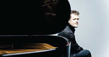 Benjamin Grosvenor, a young white man with short dark blonde, curly hair, wearing a black shirt and trousers sat behind a piano