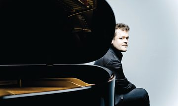 Benjamin Grosvenor, a young white man with short dark blonde, curly hair, wearing a black shirt and trousers sat behind a piano