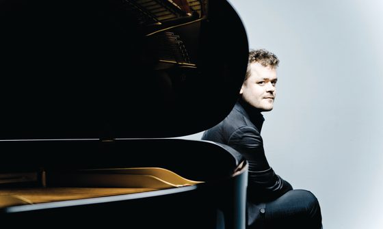 Benjamin Grosvenor, a young white man with short dark blonde, curly hair, wearing a black shirt and trousers sat behind a piano