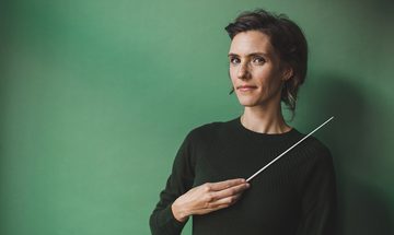 Chloe van Soeterstede, a white woman with mid length brown hair in a dark green jumper holding a conductors baton