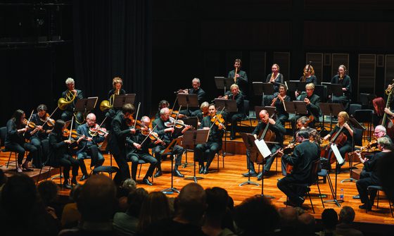 The Orchestra of the Age of Enlightenment performing on a dark stage, they're all wearing black