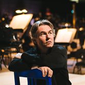 Esa-Pekka Salonen a white man with mid length floppy greying hair in a black turtle neck sitting on a blue chair backwards looking off to the right with music stands behind him
