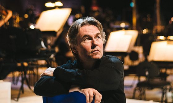 Esa-Pekka Salonen a white man with mid length floppy greying hair in a black turtle neck sitting on a blue chair backwards looking off to the right with music stands behind him