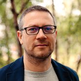 John Wilson, an older white man with short greying hair and glasses in a grey t-shirt and blue coat stood in a forest