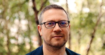 John Wilson, an older white man with short greying hair and glasses in a grey t-shirt and blue coat stood in a forest