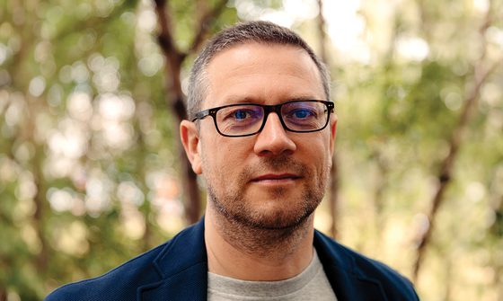 John Wilson, an older white man with short greying hair and glasses in a grey t-shirt and blue coat stood in a forest