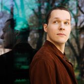 Lukas Sternath a young white man with very short brown hair in a brown suede jacket leaning against a window