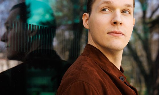 Lukas Sternath a young white man with very short brown hair in a brown suede jacket leaning against a window