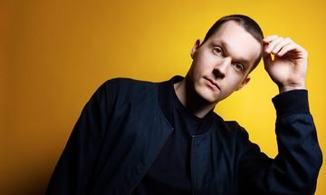 Lukas Sternath, a white man with very short brown hair leaning on his hand in a dark blue jacket on a yellow background