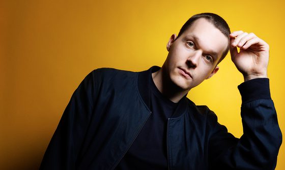 Lukas Sternath, a white man with very short brown hair leaning on his hand in a dark blue jacket on a yellow background
