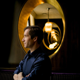 Nikolai Lugansky a white man with short brown hair in a dark blue shirt in front of a gold mirror. He's looking to the left with his arms crossed