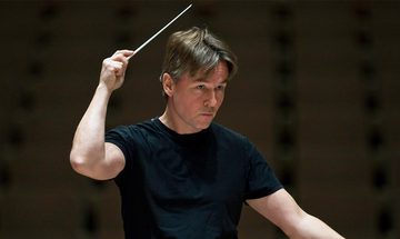 Esa-Pekka Salonen a white man with mid length floppy greying brown hair conducting in a black t-shirt with his baton above his head