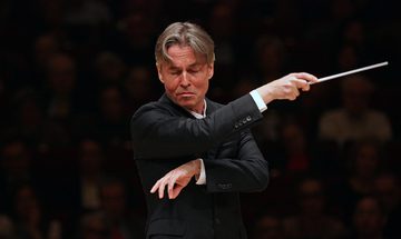 Esa-Pekka Salonen a white man with mid length floppy greying brown hair conducting in a black suit. His eyes are closed and arms crossed with his baton pointing right