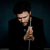 Stanislav Kochanovsky a white man with dark quaffed hair in a black suit looking down as he has his hands out in front of him in a pray motion with his baton between his hands