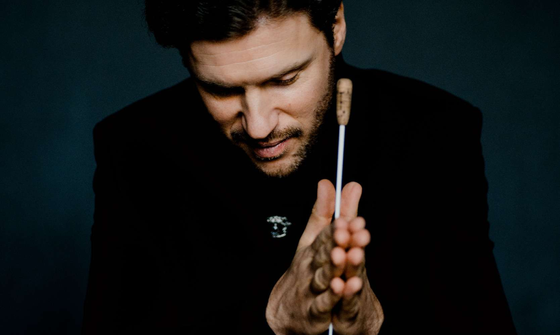 Stanislav Kochanovsky a white man with dark quaffed hair in a black suit looking down as he has his hands out in front of him in a pray motion with his baton between his hands