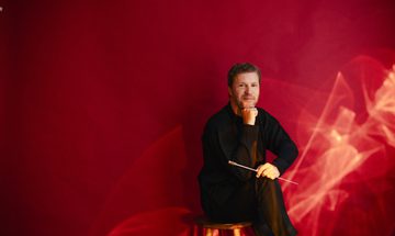 Stanislav Kochanovsky a white man with short quaffed hair and a stubbly beard in a black outfit, smiling while sitting on a stool against a red background. He has his head resting on his fist and his baton in his hand rest against his knee