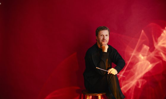 Stanislav Kochanovsky a white man with short quaffed hair and a stubbly beard in a black outfit, smiling while sitting on a stool against a red background. He has his head resting on his fist and his baton in his hand rest against his knee