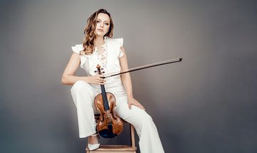Maria Ioudenitch, a young white woman with long blonde hair in a white sleeveless outfit sat on a bar stool leaning on her knee. She's holding her violin by the neck between her legs and holding the bow in the same hand